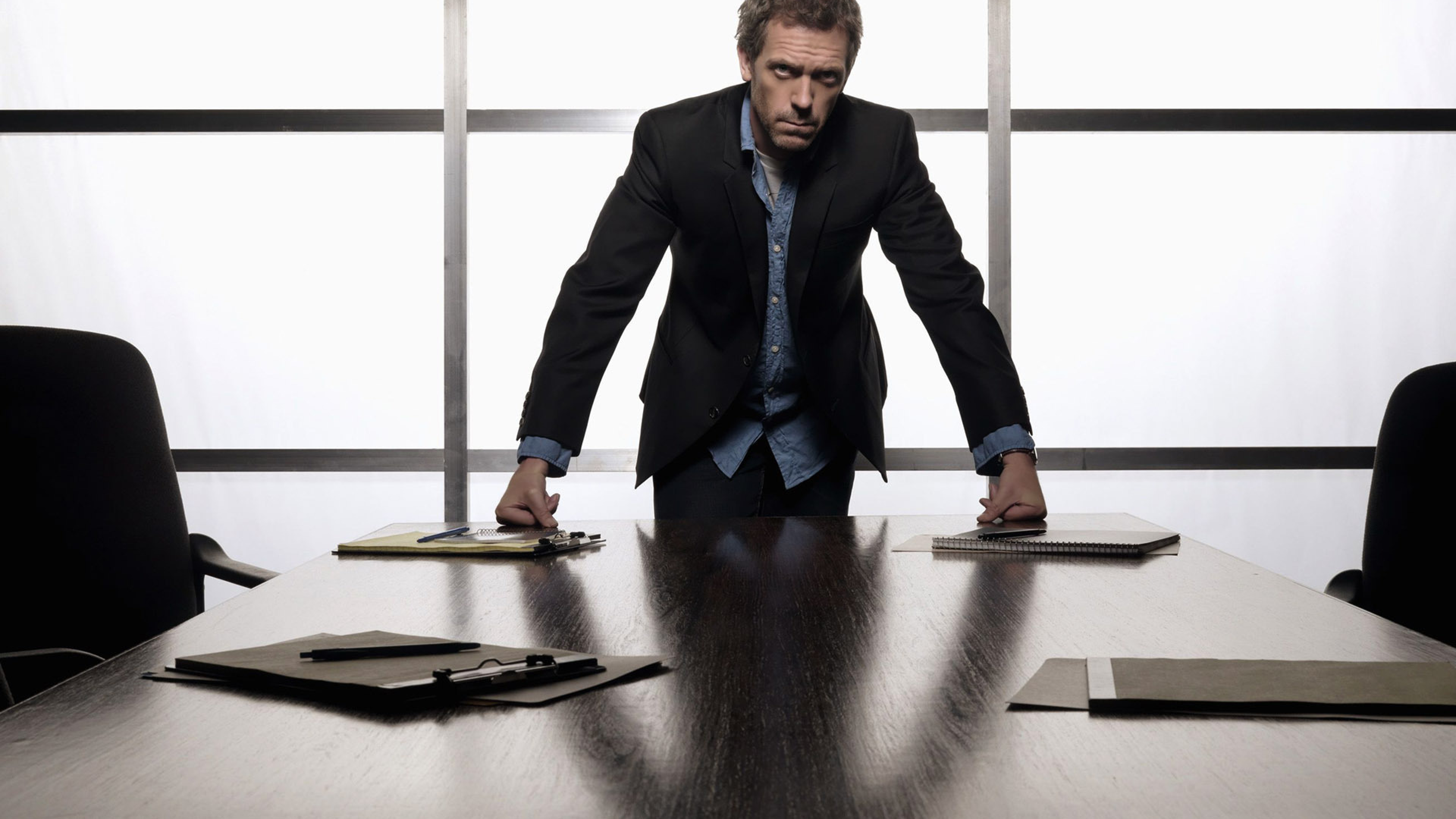 Мужчина широко сидит. Человек опирается на стол. Бизнесмен за столом. Мужчина в костюме в офисе. Властный человек.